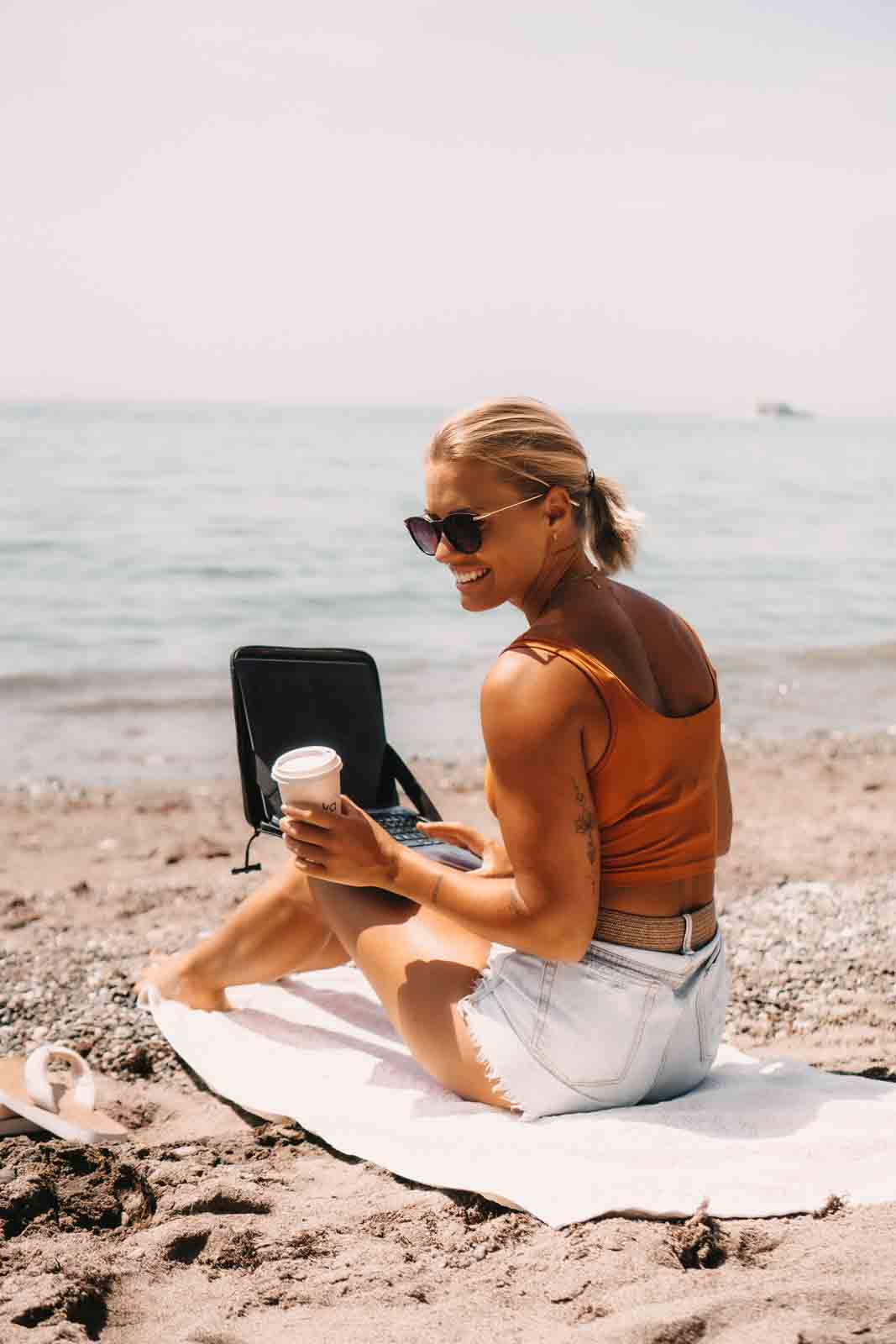 Frau am Strand mit Laptop-Tasche auf dem Schoß und Notebook als mobile Workstation – wasserdichte Laptop-Hülle für Reisen, Outdoor-Abenteuer, digitales Arbeiten und den flexiblen Arbeitsplatz am Meer.